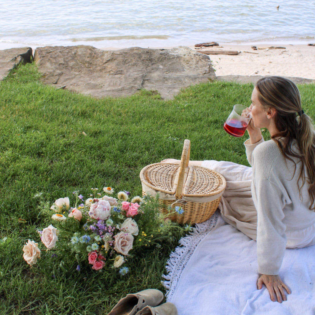 Woman enjoying a picnic by the beach with flowers and a basket.