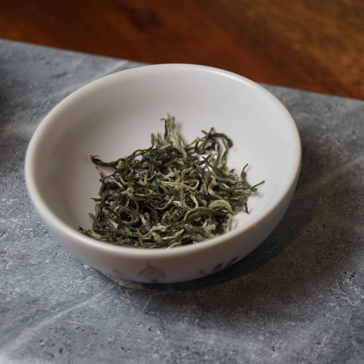 A small white ceramic bowl filled with loose green tea leaves, placed on a gray stone surface next to a white teapot with blue floral decoration.