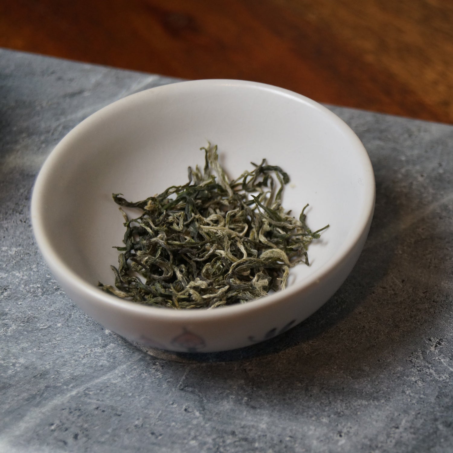 A small white ceramic bowl filled with loose green tea leaves, placed on a gray stone surface next to a white teapot with blue floral decoration.