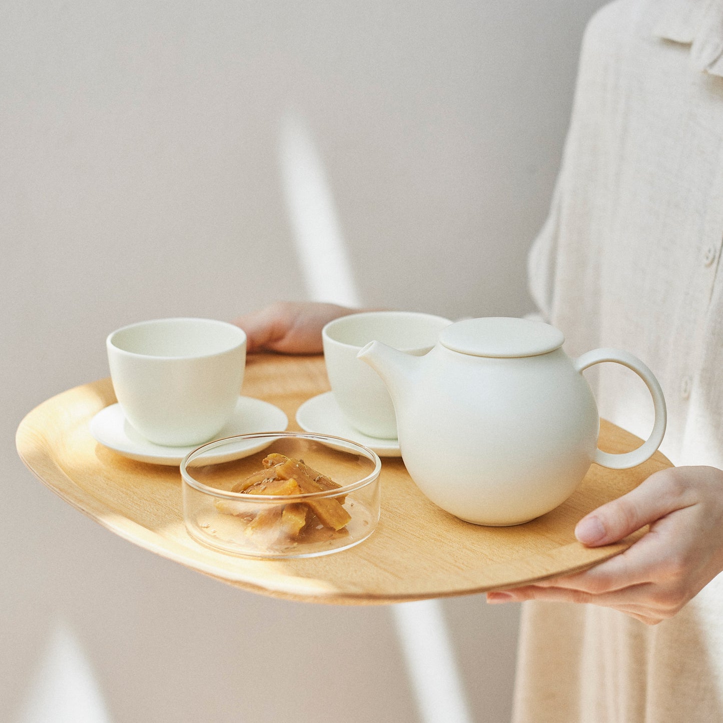 Person holding a wooden tray with ceramic tea set against a plain background