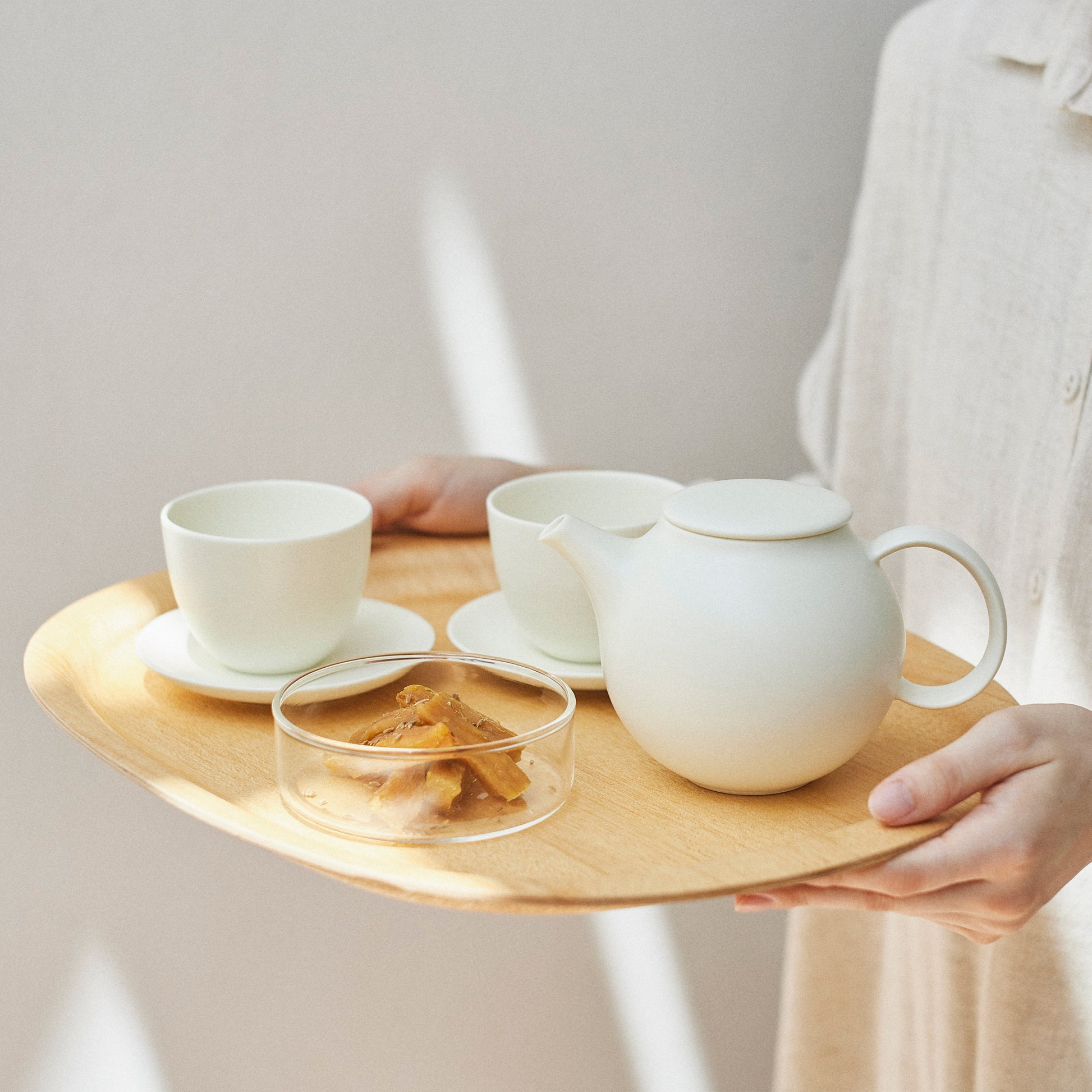 Person holding a wooden tray with ceramic tea set against a plain background