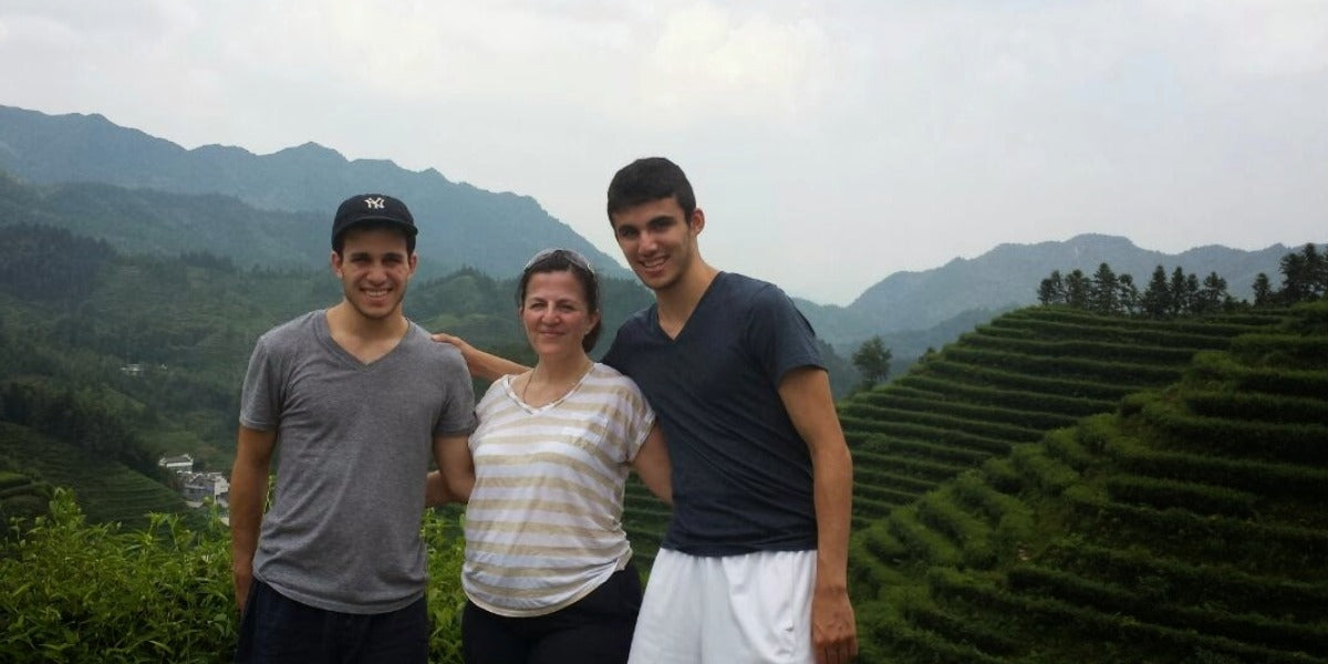 Three people standing together in front of a mountain and terraced green landscape, likely a tea plantation. The image has a casual, travel-style feel with a slightly soft, filtered quality.