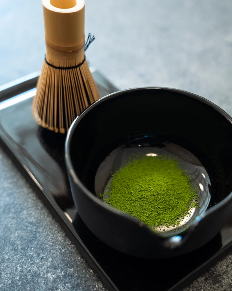 Black bowl with green tea powder on a black tray, accompanied by a bamboo whisk.
