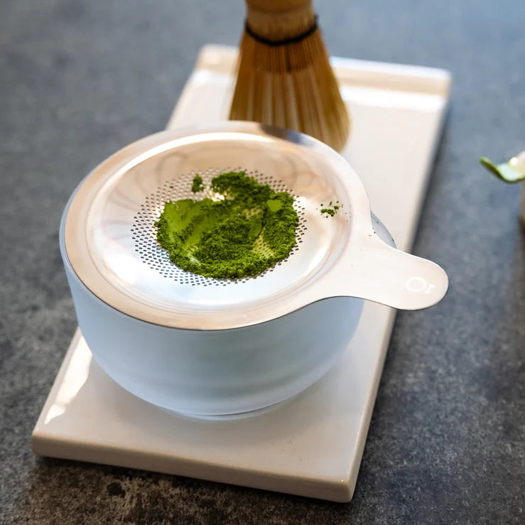 White ceramic tea strainer with green tea leaves on a gray surface