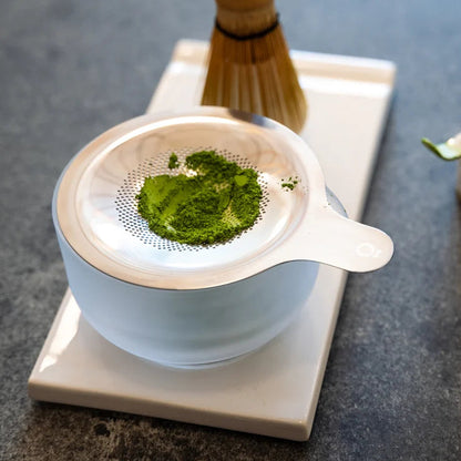 White ceramic tea strainer with green tea leaves on a gray surface